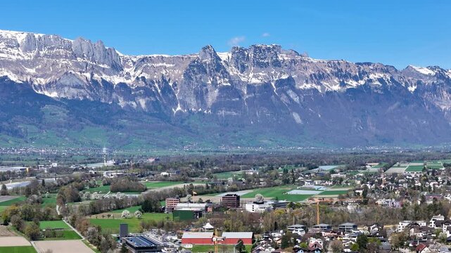 jagged alpine ridge snowcapped peaks towering rhine valley farmland below spring blossom trees small industrial buildings liechtenstein switzerland border drone footage aerial view establishing shot