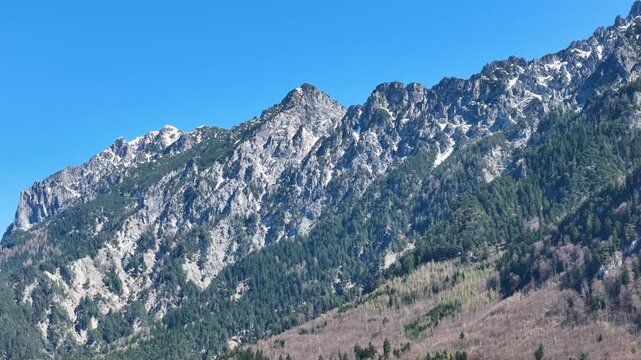 rugged mountain ridge sheer rock peaks light snow patches forested slopes clear blue sky liechtenstein switzerland border close orbit drone footage aerial view elevated view scenic landscape