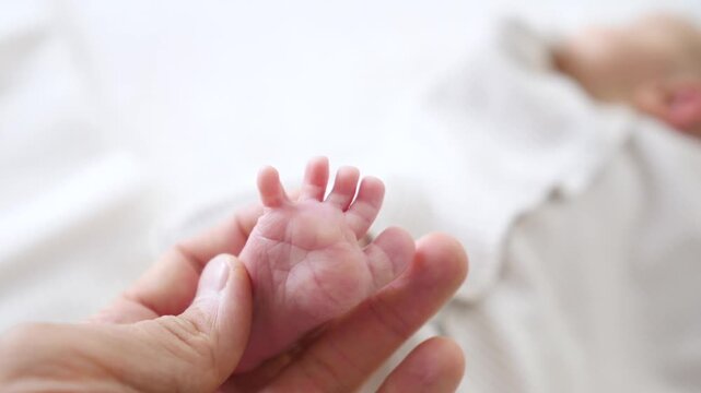 Newborn baby feet are held by adult hands in bright white bed, expressing calm tenderness in close newborn care setting. Baby feet shift slightly while fingers steady each small sole.