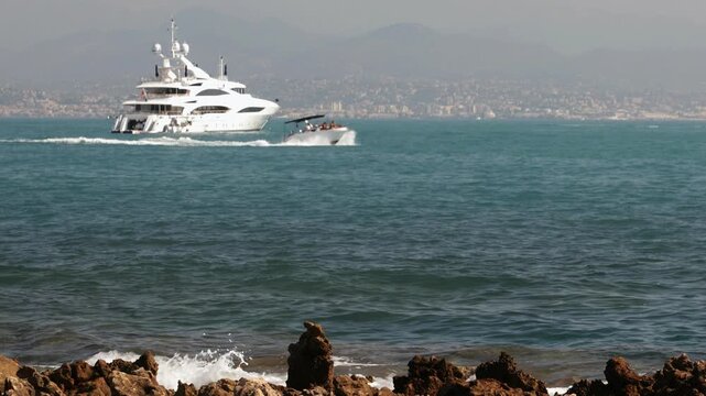 Luxury yacht anchored off Cap d'Antibes as a speedboat passes across the Mediterranean Sea, with Nice and the French Riviera visible in the background