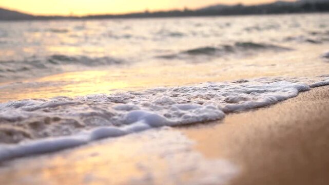 Shoreline waves wash over wet sand in golden sunset light, with tide motion rolling gently toward shore. Beach surf glows softly as water spreads and retreats, forming shifting foam along beach edge.