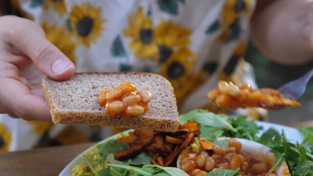 Young woman assembles bean sandwich outdoors with fork and slice of bread, smiling during vegetarian meal. Sunflower print top frames plate of roasted vegetables, greens, and baked beans on table.