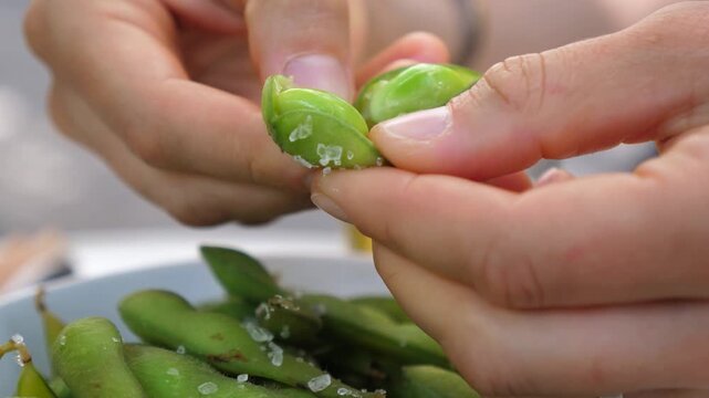 Hands shell green edamame pods in close up, calm food preparation moment with fresh vegetables and coarse salt. Hands split pods over bowl in natural light, showing careful cooking prep.