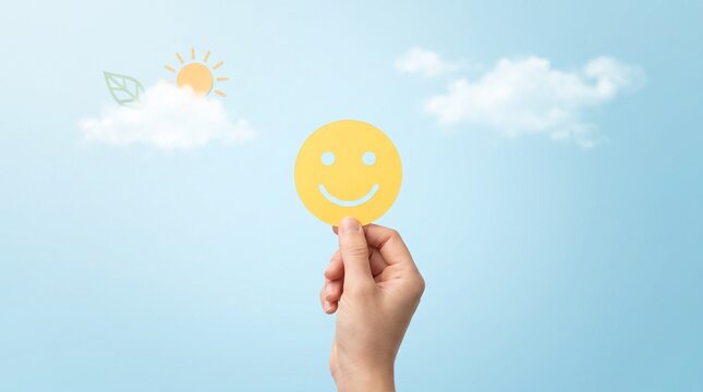 Close up of hand holding yellow smiley face symbol in front of soft blue sky with paper clouds and sun for mental wellness.