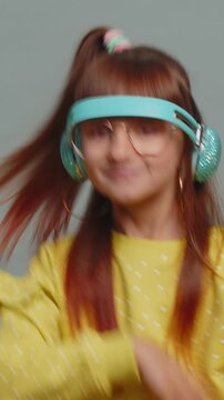 Happy preteen child girl kid listening music via headphones and dancing disco fooling around having fun expressive gesticulating hands. Little toddler children isolated on studio gray wall background.