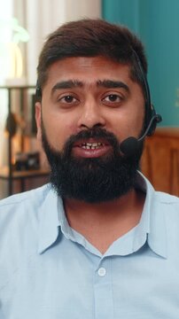Indian man at home with headset microphone speaks as help line operator explaining service to caller. Arabian guy on sofa looks into camera giving customer support guidance shows professional attitude