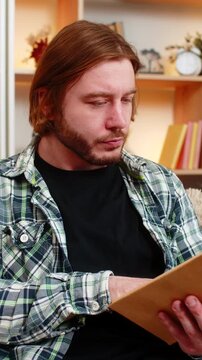 Young man on home sofa reads a few lines in book then turns page and pauses in thought. Adult boy on home sofa looks ahead for a moment while reflecting on plot meaning and ideas from reading quietly.