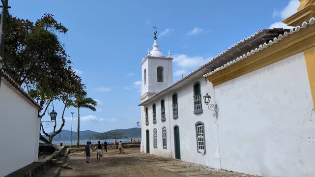 Paraty, Rio de janeiro - 2026: Slow motion static shot of the historic Capela de Nossa Senhora das Dores church next to the ocean bay, with tourists walking on the old cobblestone street.