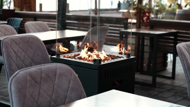 Fashionable outdoor terrace in restaurant with glass gas heater for guests comfort and relaxation, open-air patio in dining area with comfortable chairs and tables beside gas fireplace in glass box.