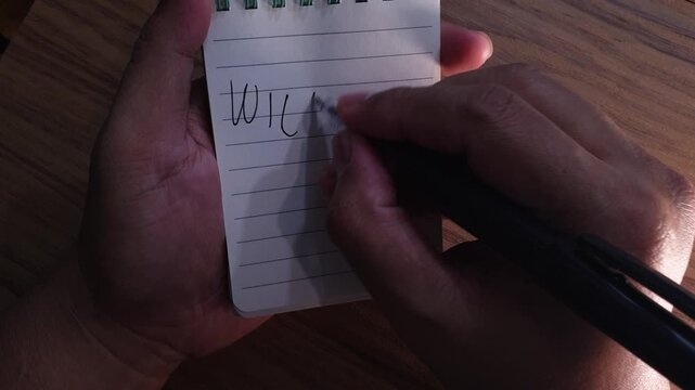 Male hands write the marriage proposal question Will You Marry Me on a small paper notepad using a black pen over a wooden desk surface.