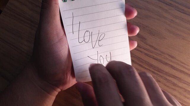 Male hands lift a lined paper note reading I Love You to reveal a hidden marriage proposal message asking Will You Marry Me on a green spiral notepad.