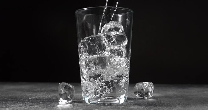 Water pouring into glass with ice cubes in slow motion on black background