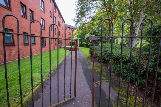 An outdoor scene featuring an old, weathered wrought iron gate that is partially open, leading into a residential area, leads into a green common area, there is a multi-story residential building. 