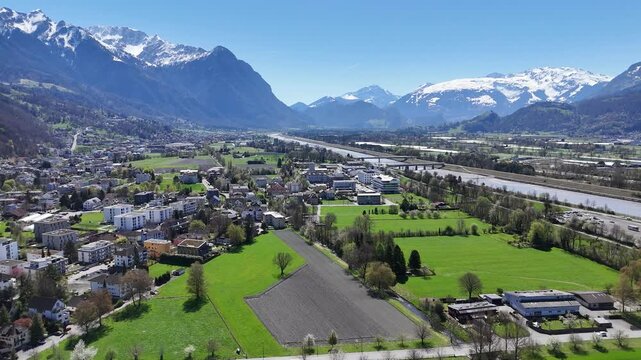 snow capped alpine peaks tower rhine valley wide panorama vaduz town rooftops foreground green farmland patchwork river gravel bed liechtenstein switzerland border aerial drone footage scenic