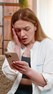 Middle-aged woman sits on home sofa reading upsetting message on smartphone fail lose online game application. Young adult girl processes bad news and loss with visible sadness and emotional stillness