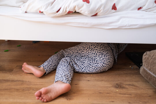 Child legs sticking out from under bed while hiding on wooden floor