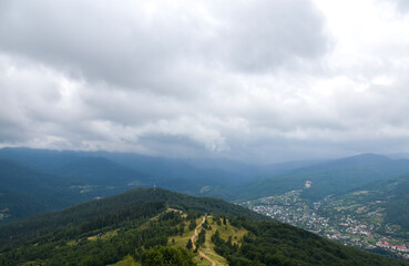 Fototapeta premium Expansive mountain landscape with rolling green hills, a winding ridge trail and a small village nestled in the valley beneath dramatic low clouds and moody overcast skies. Carpathians, Ukraine