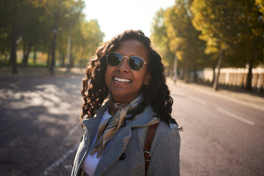 Cheerful African millennial in a gray coat and sunglasses smiles at the camera on a sunlit, tree-lined city street, with soft bokeh and autumn vibes suggesting travel and relaxed