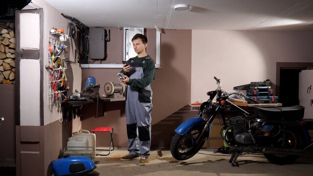 Man in overalls in a home garage holding a motorcycle engine part and checking repair instructions on his smartphone at a cluttered workbench, diy maintenance scene