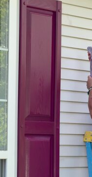 Vertical view of a hand using a cloth to wipe paint from siding and then using a paintbrush painting a house shutter red