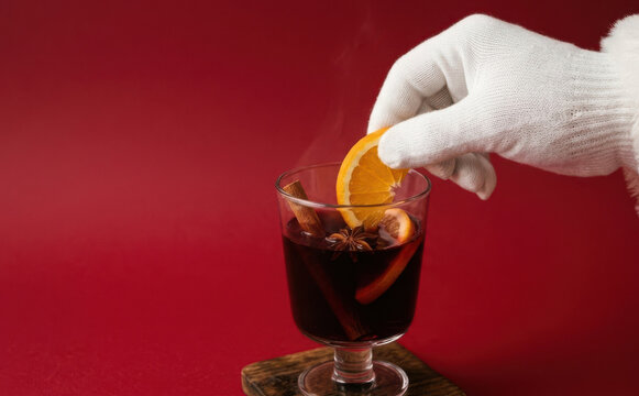 Santa hand adding orange slice to hot mulled wine with cinnamon and star anise on red background