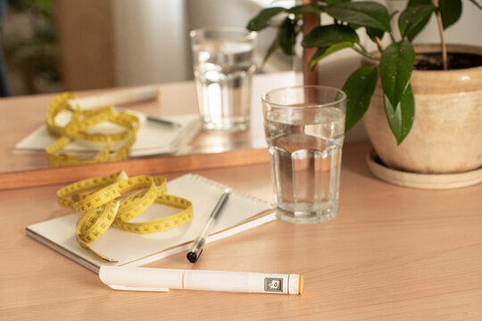 GLP-1 injection pen, measuring tape, notebook, and glass of water on wooden nightstand. Concept of medical weight loss, body weight control, health tracking, hydration, and healthy lifestyle