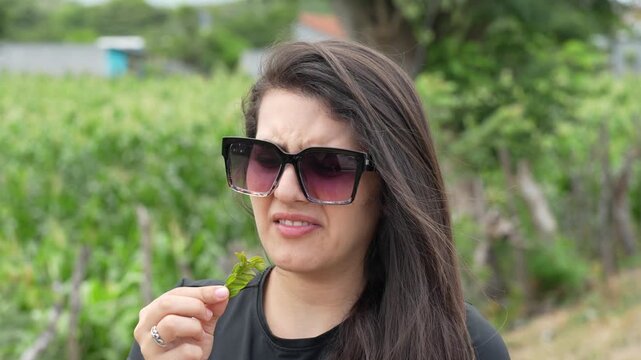 Young Woman with Sunglasses Tasting Bitter Plant Leaf and Making Disgusted Face