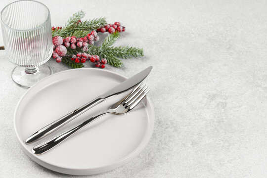 Festive holiday table setting with silver cutlery arranged on white plate with showy Christmas tree branch with red holly berry and tall glass on light gray textured background, copy space