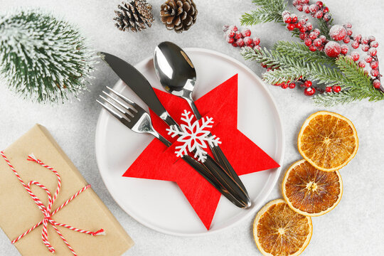 Holiday table setting with silver cutlery arranged in red star shaped cutlery holder decorated with white snowflake, Christmas tree branch, ornaments and gift box on gray textured background