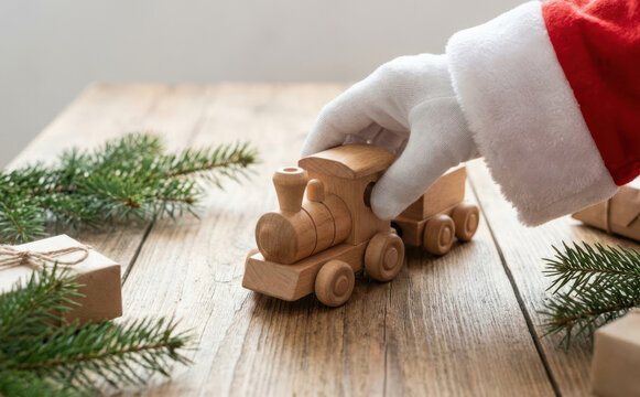 Santa Claus playing with a handmade wooden toy train on a rustic wooden table surrounded by Christmas pine branches and wrapped gifts