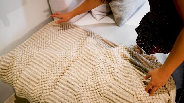 Woman hands arranging beige textured knit blanket on white sofa in modern interior.