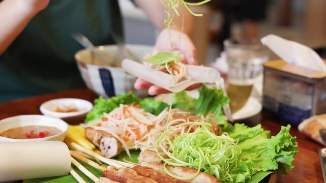 Close up of person making fresh vietnamese spring rolls with grilled pork skewers and green vegetables.