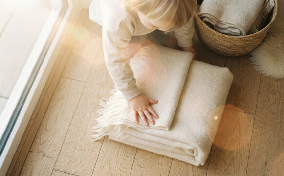 Toddling Caucasian boy folding soft wool blanket on wooden floor at home, cozy interior concept for household chores and family lifestyle photography
