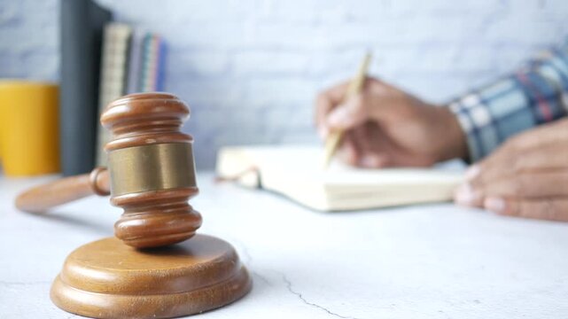 A blurred motion shot of a man's hands writing notes in a book with a pen, with a judge's gavel in sharp focus in the foreground, indicating legal study or preparation