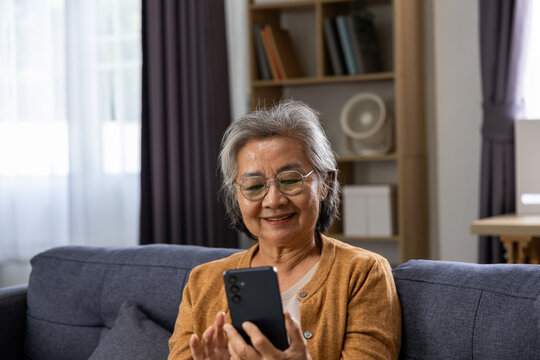 Asian senior woman using smartphone on sofa at home with warm smile and relaxed expression