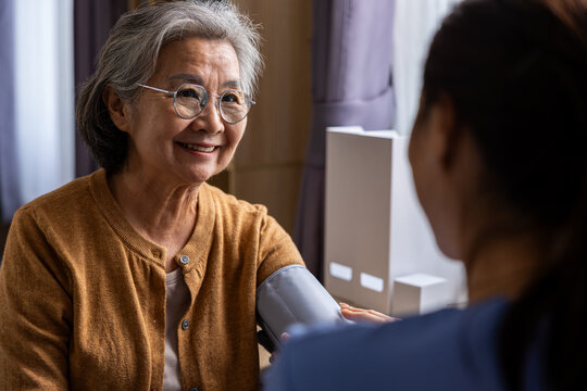 Elderly Asian woman receiving blood pressure measurement during home healthcare visit, smiling warmly at caregiver in bright room