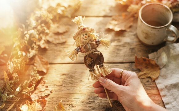 Autumn scarecrow decoration held by a woman hand on a rustic wooden table with dry leaves and a hot cup of tea for cozy fall season concept