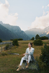 Obraz premium woman enjoying nature in mountainous meadow, sits on rock, wearing cozy cardigan and boots, peaceful outdoor lifestyle portrait with scenic landscape and natural light