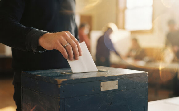 A mature caucasian man casting his vote with a ballot paper into a blue wooden box during a democratic election inside a polling station room