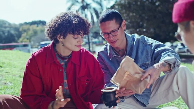 Young friends pouring dried herbal yerba mate leaves from paper craft bag into traditional calabash gourd cup, then adding hot water from thermos to brew tea during picnic on sunny day