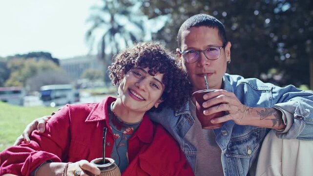 Young man and woman sitting close together on green grass in park, smiling at camera and drinking yerba mate tea from gourd cups. Video portrait