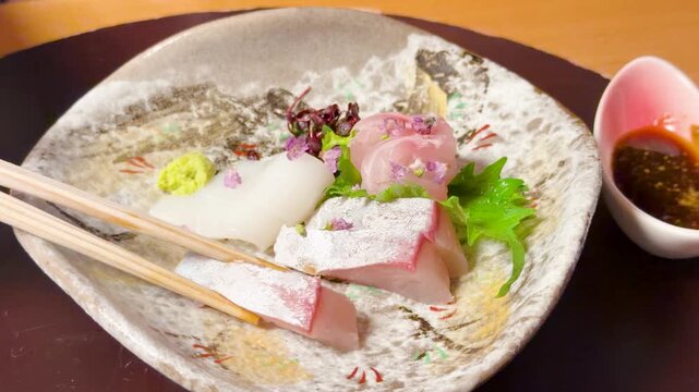 Chopsticks lifting fresh sashimi slice from ceramic plate to dip in soy sauce for traditional japanese dining experience.