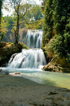 A silky waterfall pours over travertine rocks into a turquoise pool in the lush Monasterio de Piedra natural park in Aragon, Spain, a tranquil and refreshing scene of flowing water and green forest.