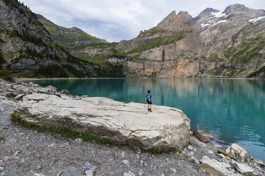 A young man in a backpack stands on a rock overlooking Oeschinen Lake (Oeschinensee) near Kandersteg, Switzerland