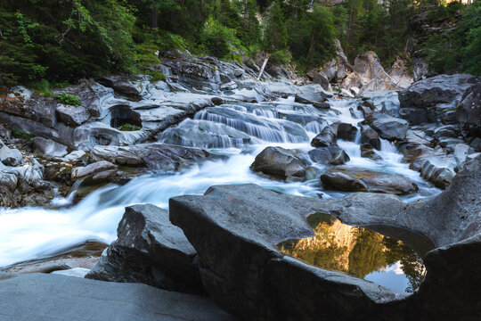 Cascades along the Avers Rhine river in the Magic Wood climbing area of Ausserferrera, Switzerland. In the foreground a small pool of water reflects golden light reflected from rock face above