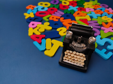 A miniature vintage black typewriter sits in front of a pile of colorful wooden letters and numbers