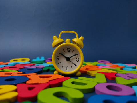 A bright yellow twin bell alarm clock stands on a blue background filled with colorful wooden letters and numbers