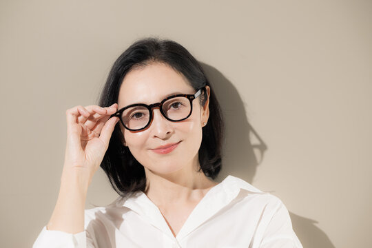 [Beige background] A close-up portrait of a woman in her 50s wearing glasses, conveying an intellectual and composed image of a middle-aged woman and her eyewear. She is looking directly at the camera