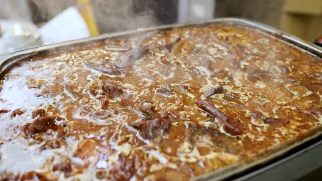 Slow cooked beef stew simmering in metal warming tray at catering event with steam rising from gravy.