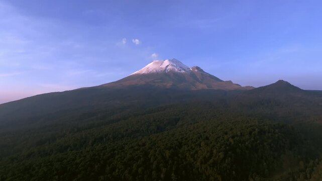 Stunning aerial view of the active Popocatepetl volcano in Mexico. The massive mountain features a snow-covered peak and a small smoke fumarole, rising above a vast, dense pine forest under a clear bl